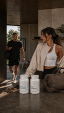 Two white supplement bottles on a stone counter in a modern indoor setting, with a woman in athletic wear standing beside them and a man walking in the background.
